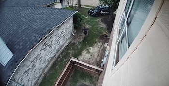 Persona corriendo por un jardín, un coche policial negro en la calle, edificaciones residenciales con tejados oscuros y una valla de madera