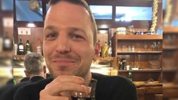 Primer plano de un hombre sonriente con barba incipiente, sosteniendo un pequeño vaso de licor, con un bar o estantería de restaurante al fondo