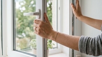 Primer plano de dos antebrazos y manos abriendo una ventana blanca. Se ven un picaporte, un anillo, una pulsera, cortinas claras y vegetación exterior