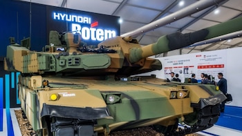 Tanque militar camuflado con logo "Hyundai Rotem" en exhibición. Varias personas observan un panel informativo al fondo