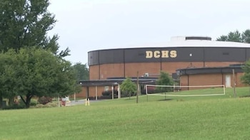 Vista exterior del edificio de la DCHS, con paredes de ladrillo y una sección superior oscura, frente a un campo de hierba verde con una cancha de voleibol y árboles
