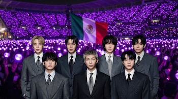 Siete miembros de BTS en trajes oscuros posan frente a un estadio con luces violetas y la bandera de México.