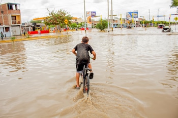 Un niño avanza de espaldas en bicicleta sobre una calle cubierta de agua marrón frente a postes y edificaciones urbanas