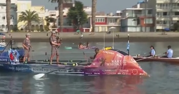 Dos hombres en un bote azul y blanco, uno toca la gaita. Otro bote rojo y un bote de remo con dos personas están en el agua. Palmeras y edificios en el fondo