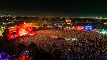 Vista aérea nocturna de un festival con una multitud masiva. Múltiples escenarios brillantemente iluminados con luces rojas, azules y verdes