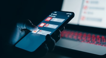 Primer plano de una mano sosteniendo un teléfono inteligente en una habitación oscura con luz azul; al fondo, un teclado de laptop con brillo rojo.