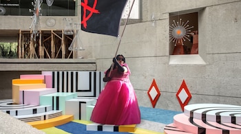 Persona con vestido rosa y gafas de sol sostiene una bandera negra con un símbolo anarquista rojo ante estructuras de colores.