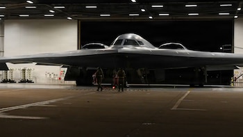 Vista frontal de un bombardero B-2 Spirit gris oscuro dentro de un hangar. Dos militares con equipo de tierra están de pie cerca de la parte delantera del avión