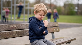 Niño rubio de piel clara sentado en un banco de madera en un patio. Viste sudadera azul marino y pantalón gris. Fondo borroso con juegos y niños.