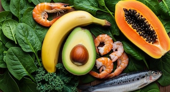 Vista aérea de diversos alimentos frescos sobre hojas verdes: un plátano, una palta partida, una papaya partida, camarones y una sardina.