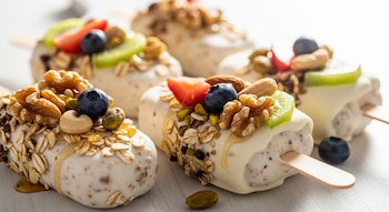 Varias paletas de yogur y avena en primer plano con cobertura blanca, decoradas con arándanos, fresas, kiwi, nueces, anacardos, pistachos y avena sobre un fondo claro.