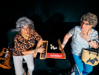 Vista aérea de dos mujeres mayores con atuendos elegantes jugando Rummikub en una mesa negra con fichas visibles. Una mira a la cámara, la otra con expresión de preocupación