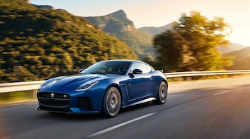 Jaguar F-Type coupé azul en la carretera. Montañas cubiertas de árboles y un cielo con colores de atardecer en el fondo, con efecto de movimiento.