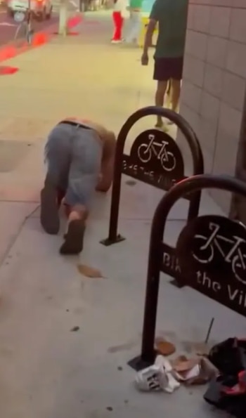 Hombre arrodillado en acera. Viste pantalones cortos y calzado oscuro. Hay dos portabicicletas negros y basura en el suelo. Personas y calle en segundo plano