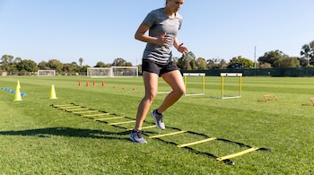 Mujer de camiseta gris y pantalones cortos negros pisa una escalera de agilidad amarilla en un campo verde con conos y vallas de entrenamiento.