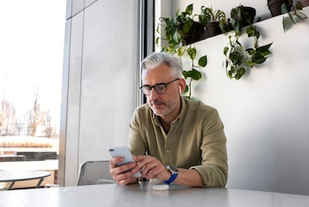Un hombre con gafas y cabello canoso sostiene un teléfono inteligente sentado ante una mesa con plantas de fondo