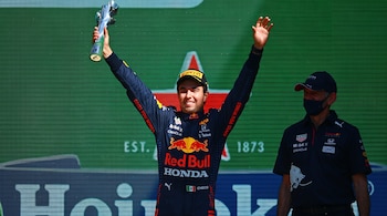 Sergio Pérez con traje deportivo de Red Bull sostiene un trofeo plateado con los brazos en alto junto a un integrante del equipo.