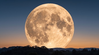 Gran Luna llena de color anaranjado sobre una silueta oscura de árboles en la base, bajo un cielo con degradado azul y naranja.