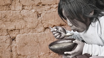 Una niña con pelo largo y oscuro, vestida de claro, come de un cuenco con una cuchara, con una pared de adobe de fondo marrón rojizo.