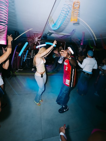 Personas bailando en una fiesta nocturna. Un hombre con camiseta roja, una mujer de pelo rubio. Techo con espirales de colores y efectos de luz