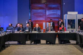 Grupo de personas sentadas en una larga mesa con micrófonos y vasos de agua. Hombres con traje y una mujer con chaqueta roja. Al fondo, personas de pie y dos puertas de madera
