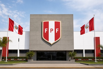 Fachada de un edificio gris y blanco con el escudo de la Federación Peruana de Fútbol en el centro y dos banderas peruanas a los lados.