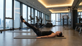 Mujer tumbada boca arriba en esterilla gris elevando las piernas en un gimnasio moderno. Viste top y leggings negros, zapatillas oscuras. Grandes ventanales y equipos de fitness al fondo.