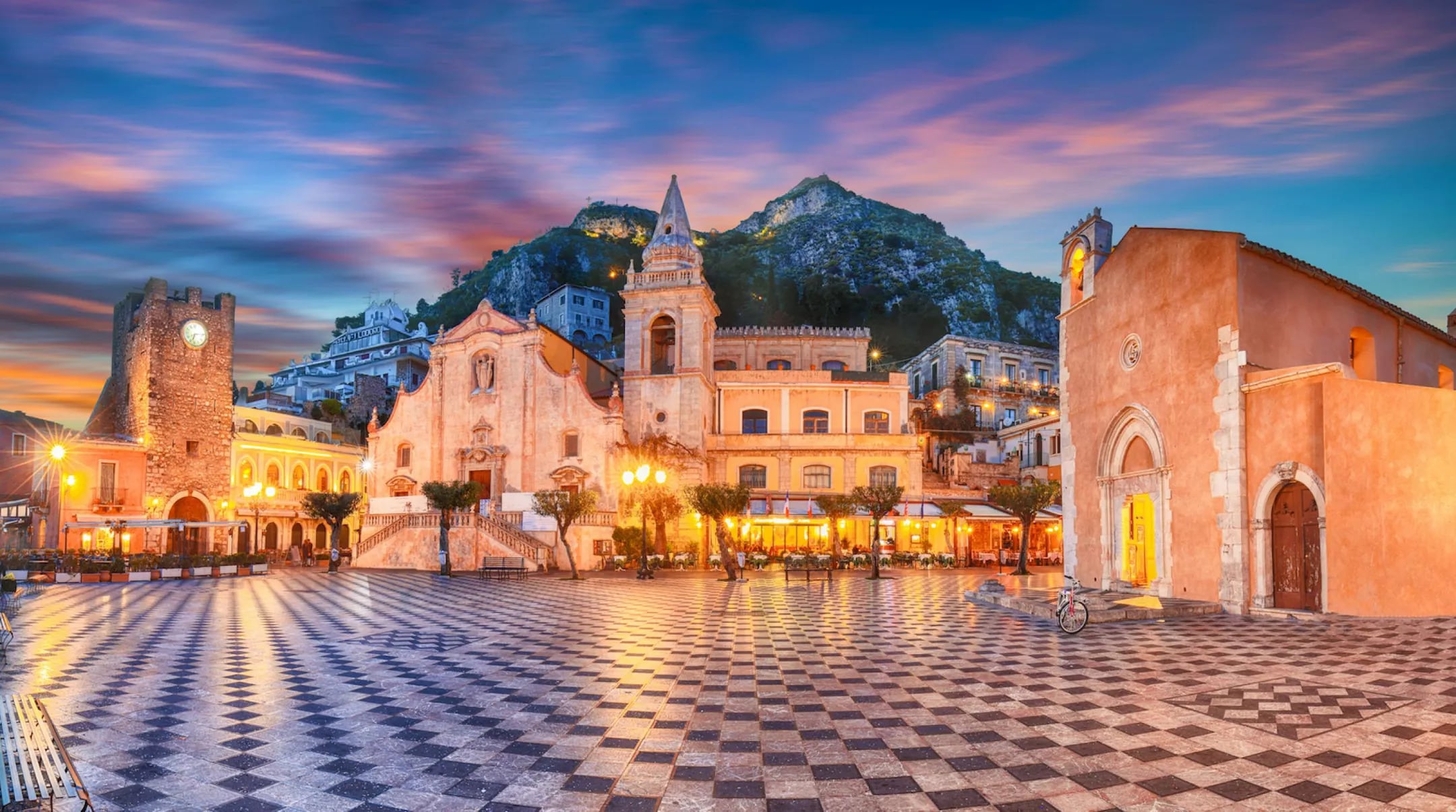 La Piazza IX Aprile en Taormina reúne edificios históricos como la iglesia de San José y la torre del Reloj frente a su pavimento ajedrezado iluminado por farolas. (Ministerio del Turismo de Italia)