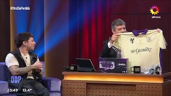 Mario Pergolini sostiene una camiseta beige frente a Frankkaster en un estudio de televisión con un escritorio y un teclado en caja