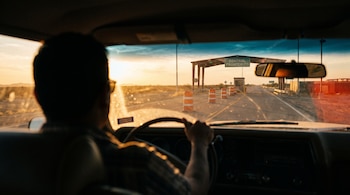 Vista desde el interior de un automóvil hacia un puesto de control fronterizo con conos de tránsito en una carretera al atardecer.