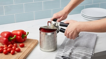 Primer plano de unas manos usando un abrelatas para abrir una lata de salsa de tomate en una encimera blanca con azulejos celestes. Pimientos y tomates cherry cerca.