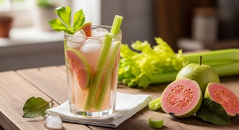 Vaso de agua de guayaba y apio con hielo, rodajas de guayaba y tallos de apio en una mesa de madera. Guayabas frescas enteras y cortadas al lado.