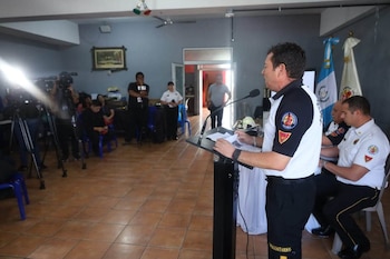 Hombre de uniforme de bombero habla en atril. Sentados, otros dos hombres de uniforme. Al fondo, público, cámaras de video, banderas nacionales y un casco