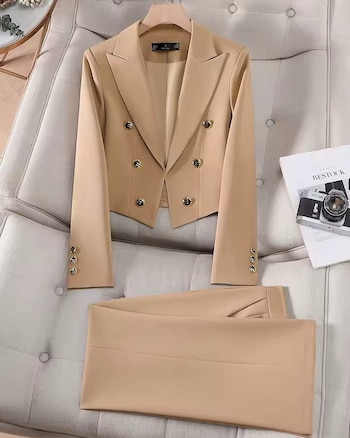 Un traje de dos piezas color beige, que incluye un blazer corto con seis botones dorados y pantalones, colocado sobre un sofá tapizado claro