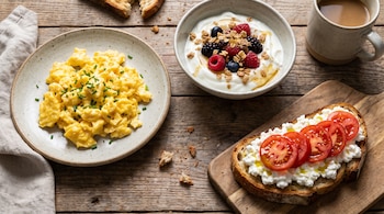 Huevos revueltos, un tazón de yogur griego con bayas y granola, una tostada con queso cottage y rodajas de tomate, y una taza de café sobre una mesa de madera.