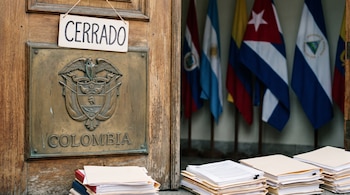Puerta de madera con placa de bronce del escudo de Colombia, cartel de 'Cerrado', papeles y carpetas apilados, y banderas desenfocadas de fondo.