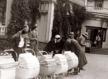 Imagen en blanco y negro de un grupo de adultos, hombres y mujeres, mirando múltiples cochecitos de bebé frente a una edificación con hiedra