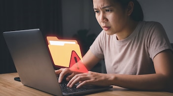 Persona teclea en un computador portátil con el ícono de una carpeta y una señal de advertencia en la pantalla, las manos están sobre el teclado.