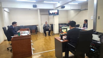 Una sala de audiencia con Daniel Algarilla sentado en el centro hablando a un micrófono. Otros hombres están sentados en mesas. Una pantalla en la pared muestra una videollamada