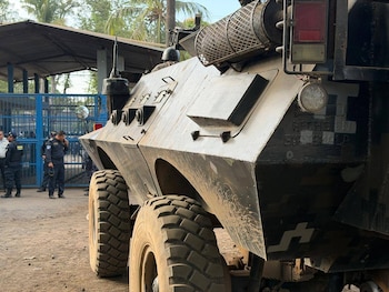 Primer plano de un vehículo blindado oscuro con ruedas grandes, mostrando "Hecho en Guatemala" en el lateral. Policías y una valla azul en el fondo