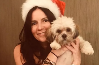 Una mujer de cabello oscuro y largo, con un gorro de Papá Noel, sonríe mientras sostiene a un perro pequeño de pelaje blanco y marrón claro