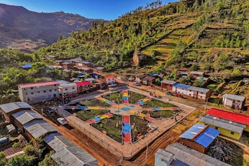 Vista aérea de un pueblo rural peruano en un valle andino, con un parque central de senderos geométricos coloridos rodeado de casas sencillas