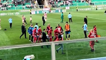 En un campo de fútbol, personal médico y de emergencia con uniformes rojos y negros retiran a un jugador en camilla, rodeados de otros individuos
