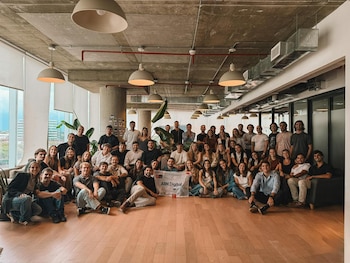 Una gran audiencia de unas 50 personas posa sonriendo en una oficina moderna con techo de hormigón, lámparas colgantes y piso de madera