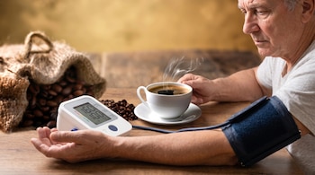 Un hombre sentado junto a un tensiómetro digital en su brazo, una taza de café humeante y un saco con granos de café.