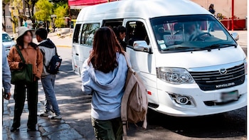 Tres personas, dos de ellas con mochilas, se encuentran en una acera junto a una camioneta tipo van blanca de marca King Long en una calle