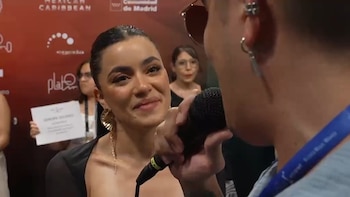Una mujer con cabello oscuro recogido y aretes dorados sonríe mientras sostiene un micrófono negro durante una entrevista, con logos de evento de fondo