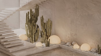Interior contemporáneo con escalera flotante de piedra clara, grandes cactus en jardineras con guijarros blancos y luces esféricas, y paredes texturizadas beige.