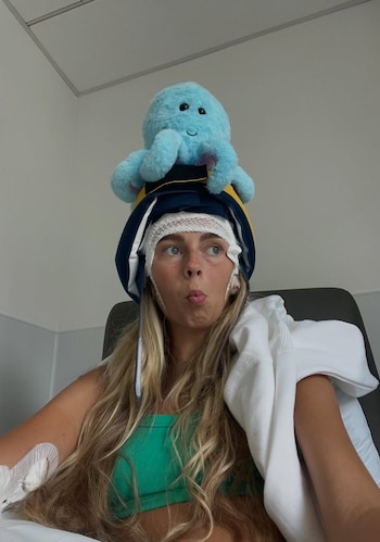 Mujer joven rubia con vendajes en la cabeza, gorro de graduación azul y amarillo, peluche de pulpo azul, y una vía en el brazo