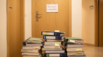Varias pilas de carpetas y libros con notas adhesivas de colores apilados en el suelo frente a una puerta de madera. Un letrero en la puerta dice 'Revisión curricular'.